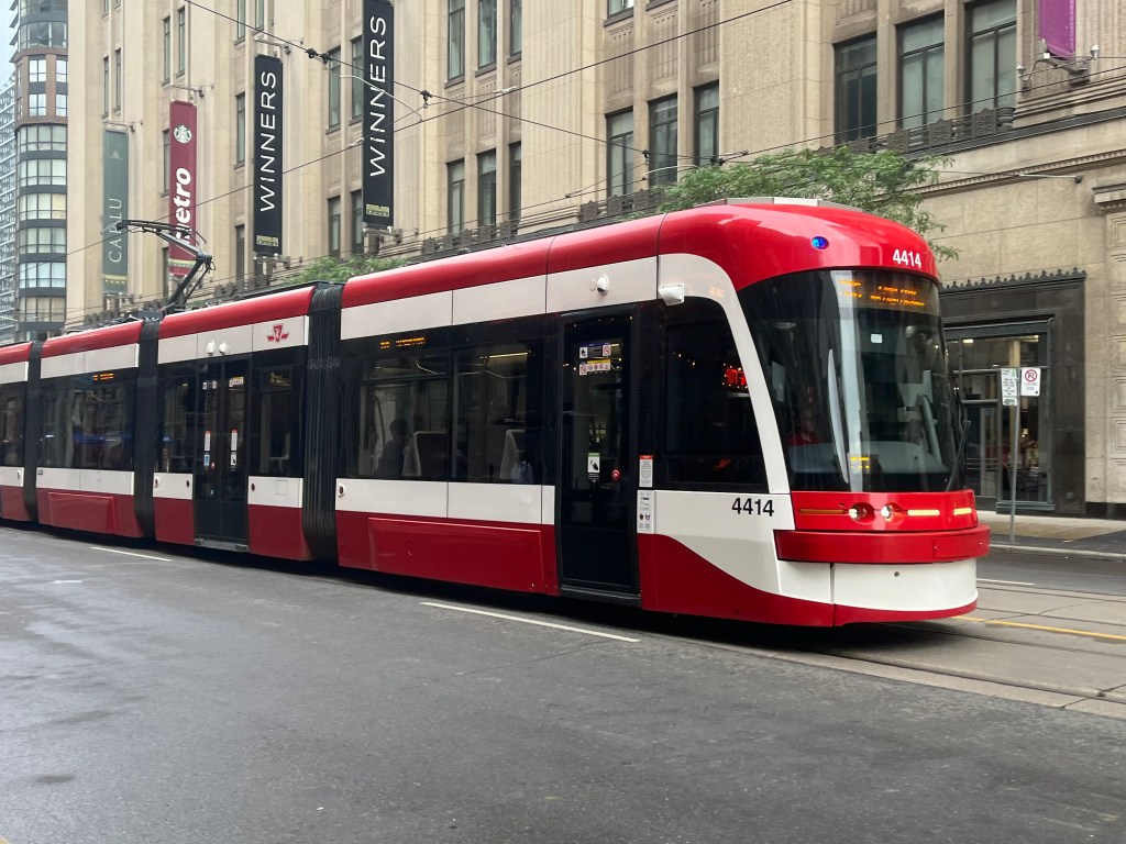 A red and white slim,  long street car in the middle of the street, with grey concrete building in the background 